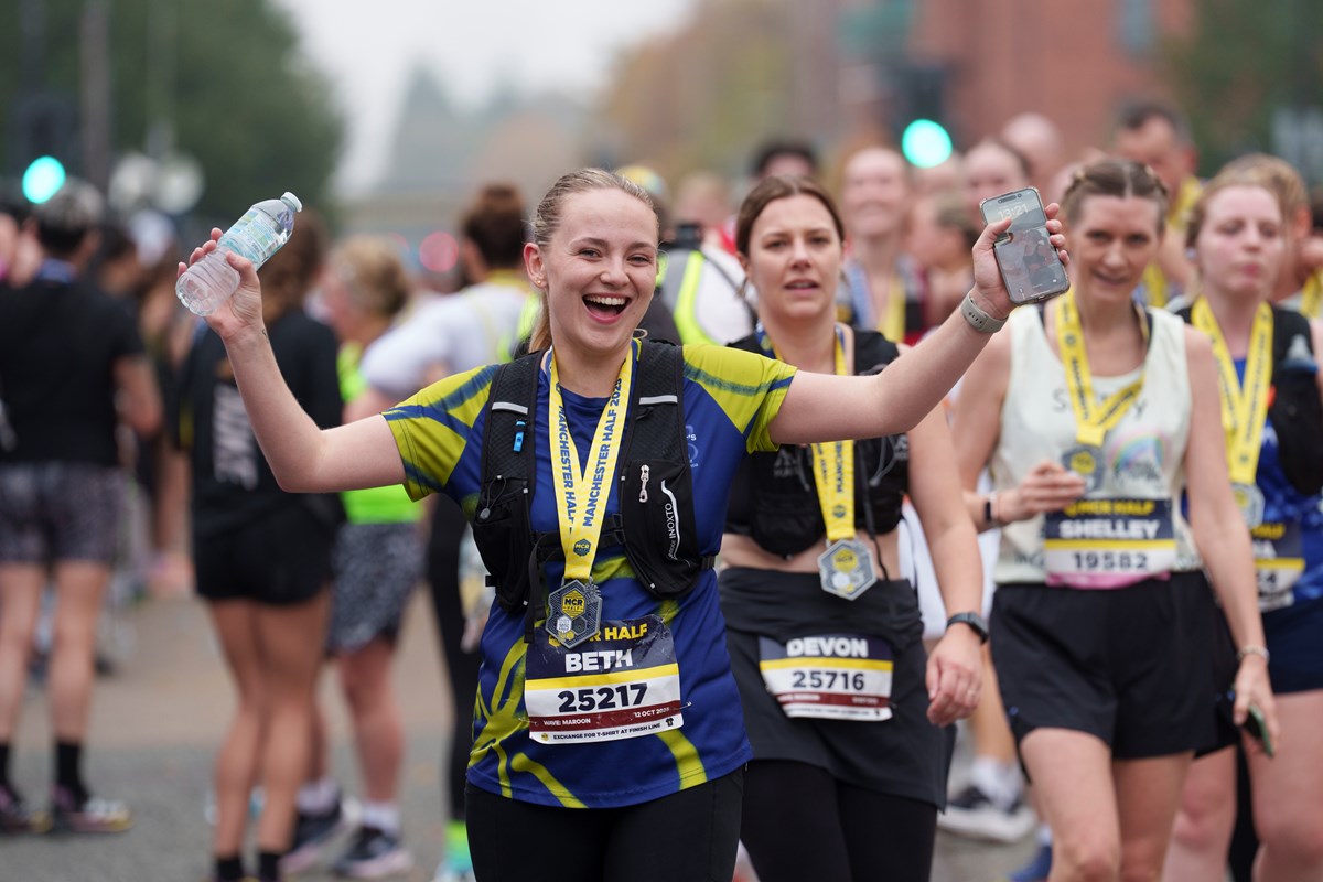 A smiling runner raises her arms in celebration after finishing the Manchester Half, wearing a finisher's medal, with other runners behind her.