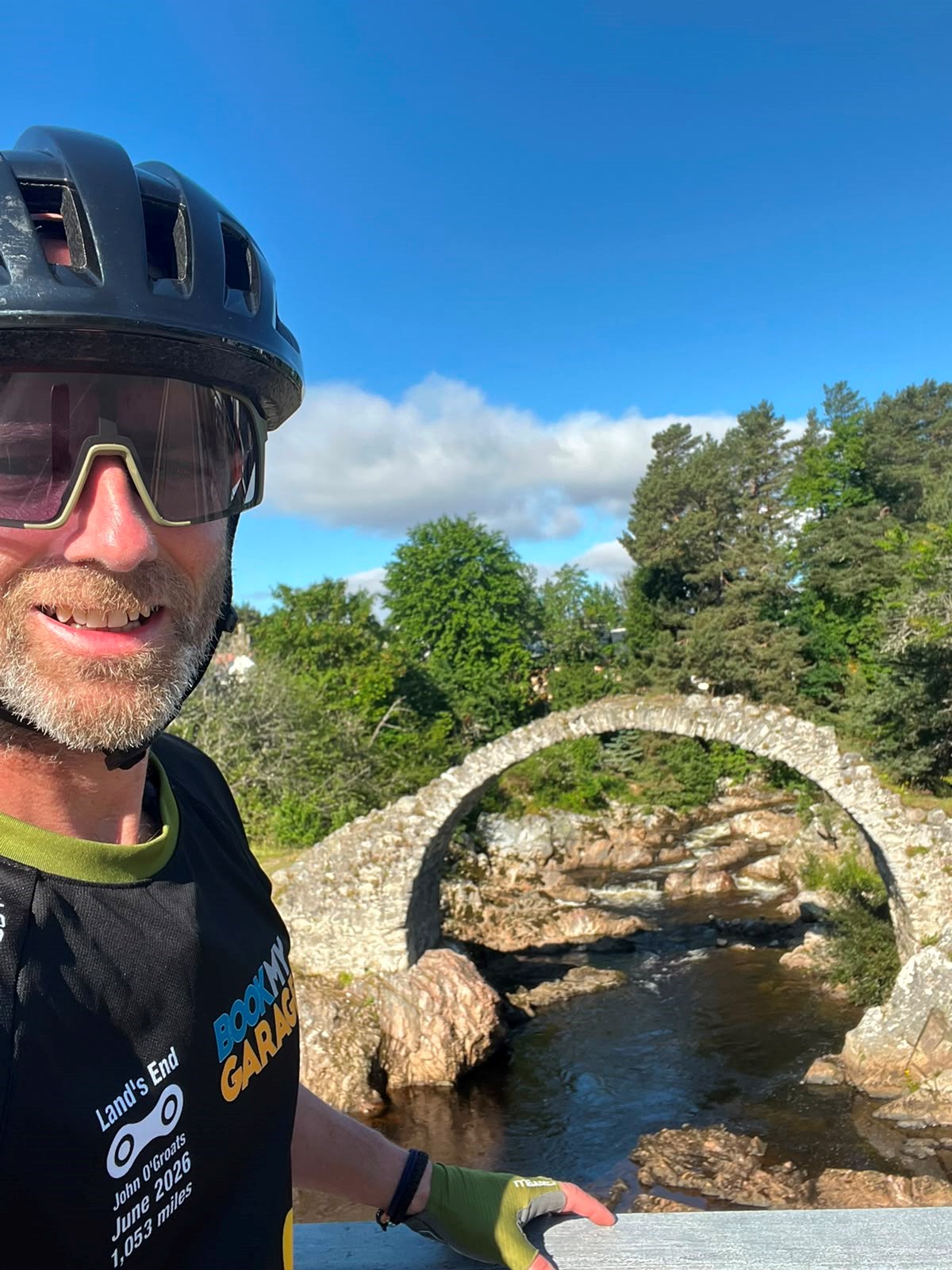 Cyclist in helmet and sunglasses taking a selfie in front of an old stone arched bridge over a river, wearing a jersey reading "Land's End to John O'Groats, June 2026, 1,053 miles".