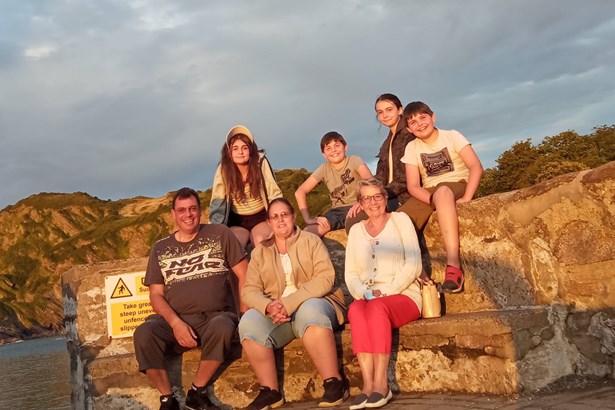 A family of seven, three adults and four children, sitting together on a stone harbour wall by the sea at sunset.