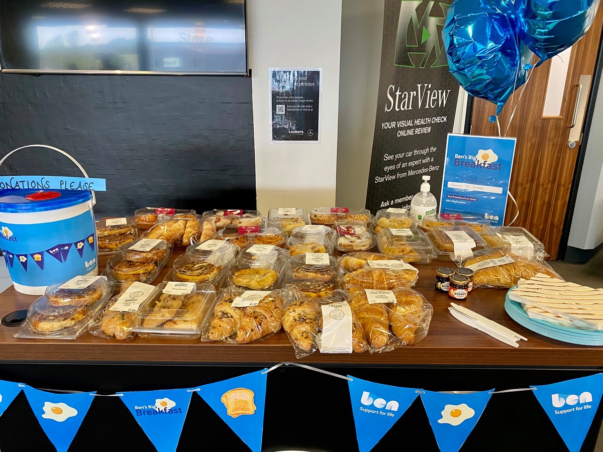 A Ben's Big Breakfast fundraising table in a car dealership, laid with packaged pastries, a donations bucket, blue balloons and Ben bunting.