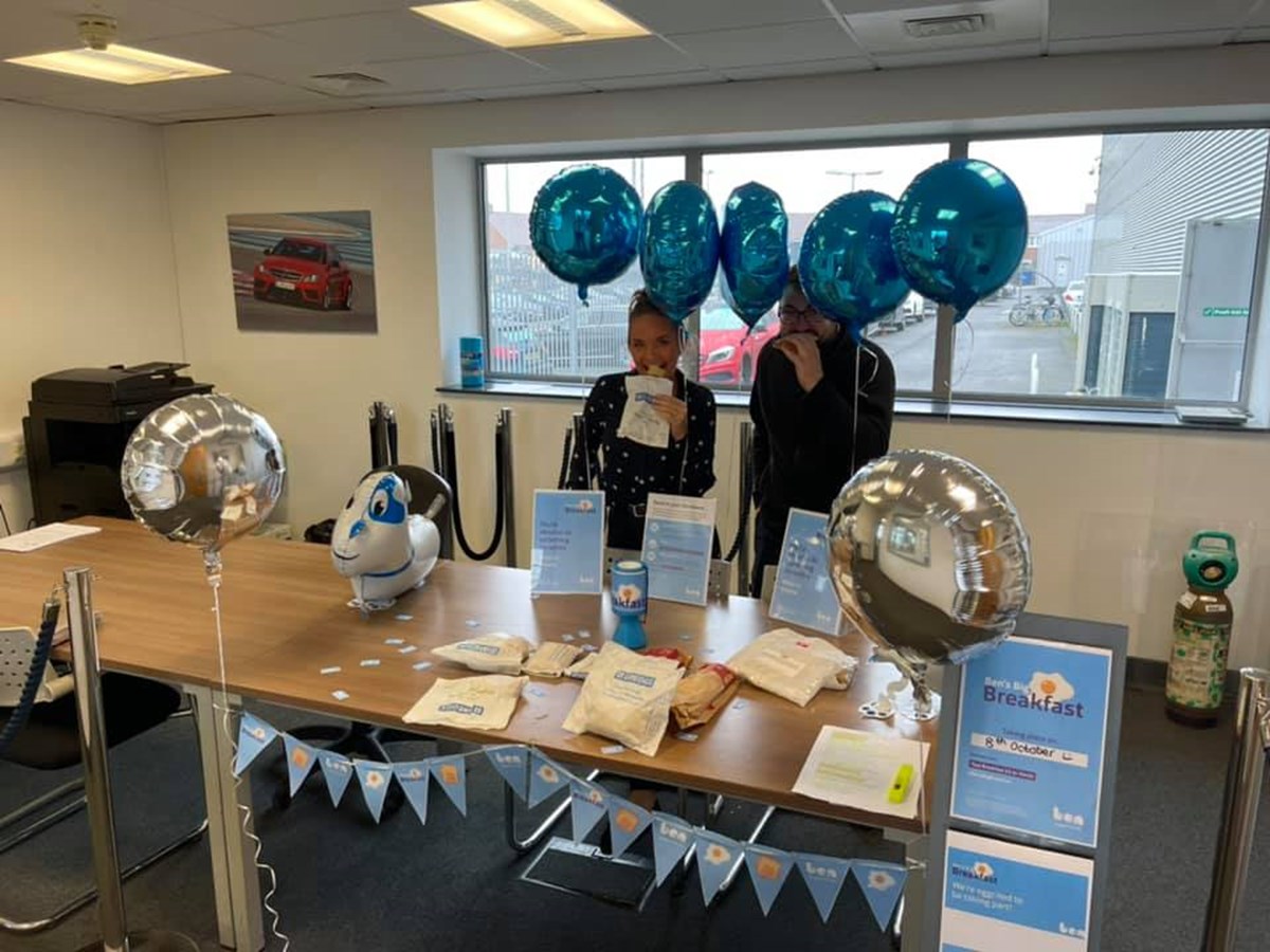 Two colleagues eating breakfast rolls behind a table decorated with Ben's Big Breakfast bunting, balloons and a collection tin.
