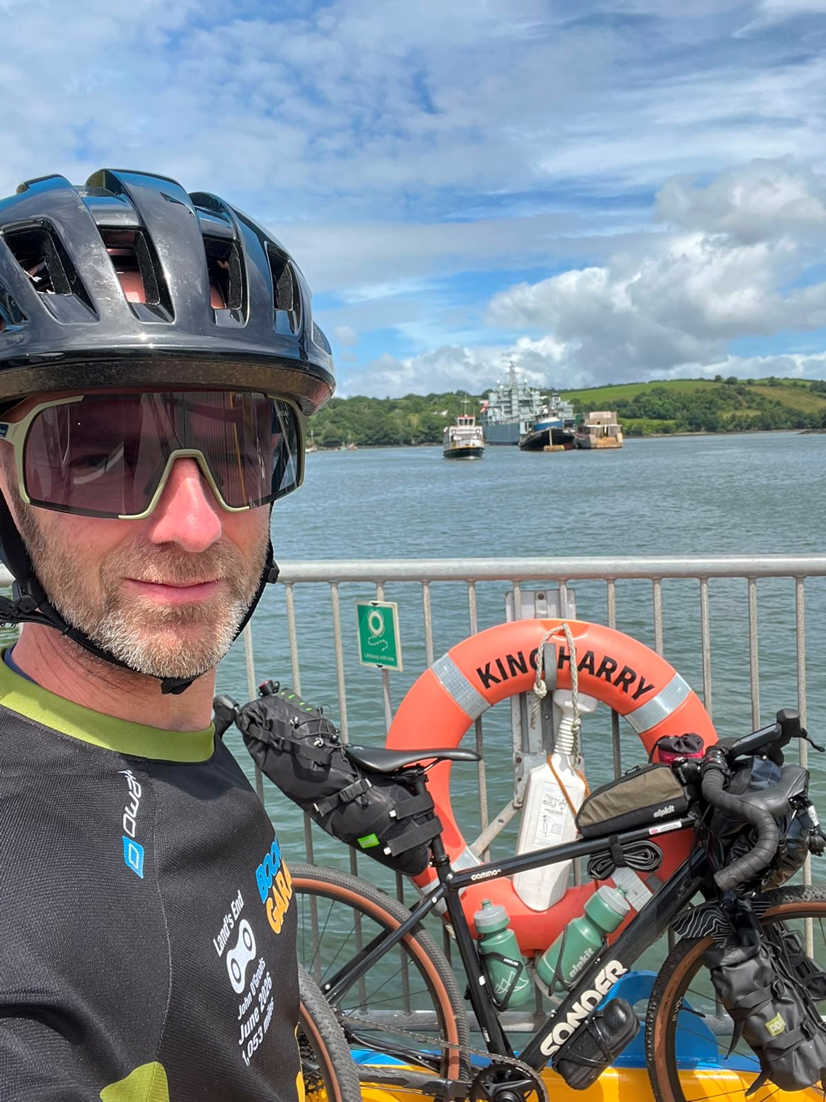 Cyclist in helmet and sunglasses takes a selfie on a river ferry, with his loaded bike beside a 'King Harry' lifebuoy.