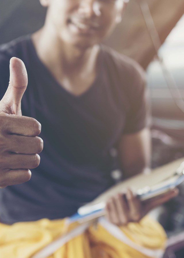 A person perched on an open car bonnet in a workshop smiling doing a thumbs up sign