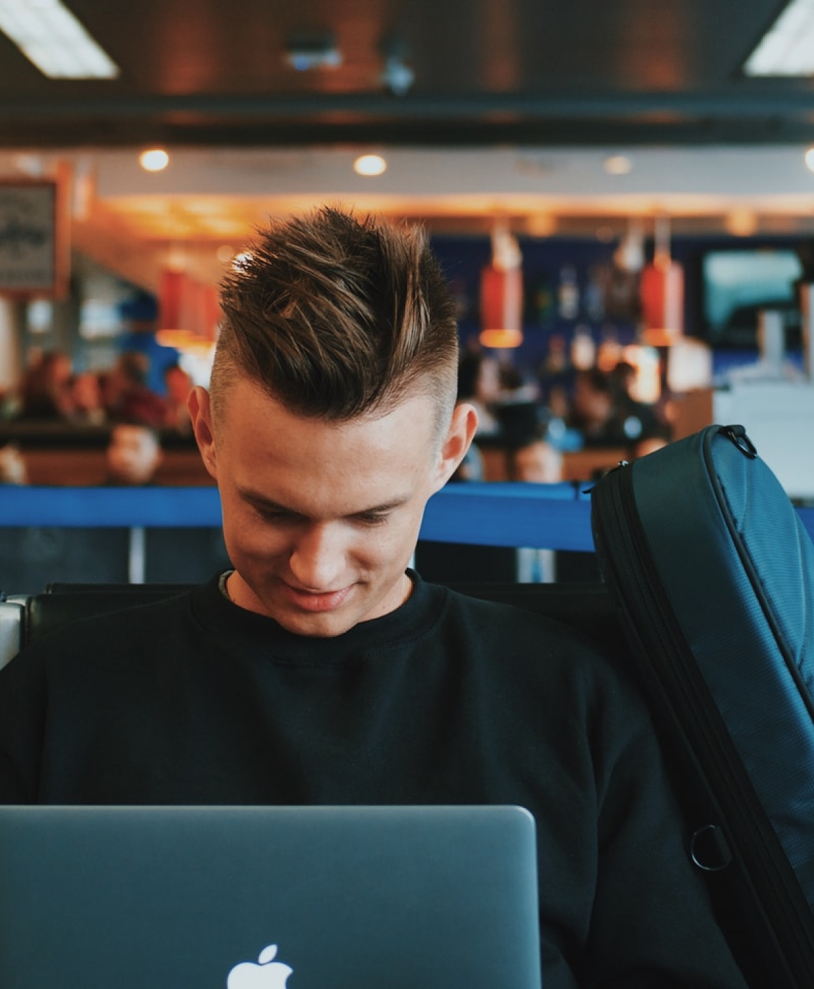 A young man smiling as he works on a laptop in a busy café, a rucksack beside him.