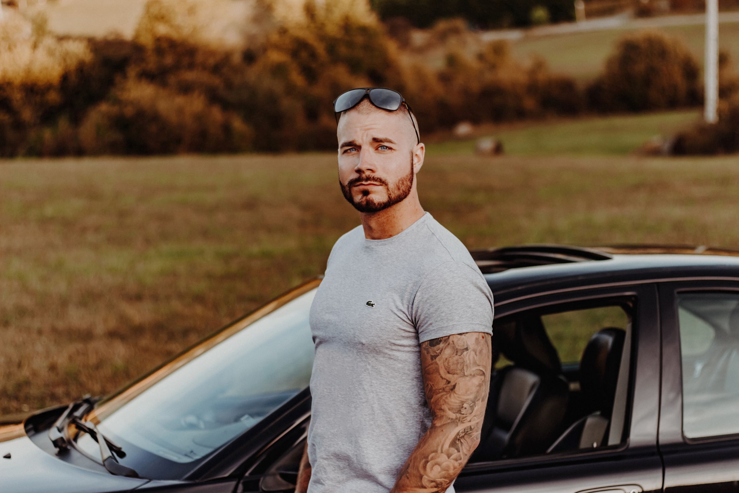 Man with sunglasses on top of his head with tattooed arm standing next to a car staring at the camera with a concerned look on his face.