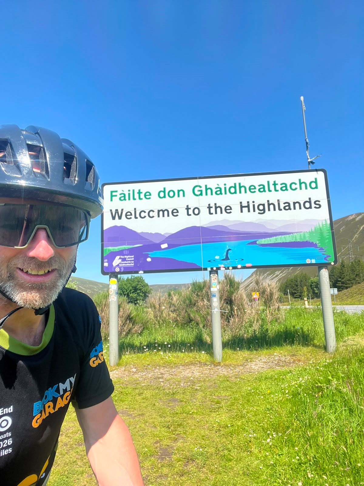Cyclist in helmet and sunglasses taking a selfie beside a road sign reading "Fàilte don Ghàidhealtachd – Welcome to the Highlands".