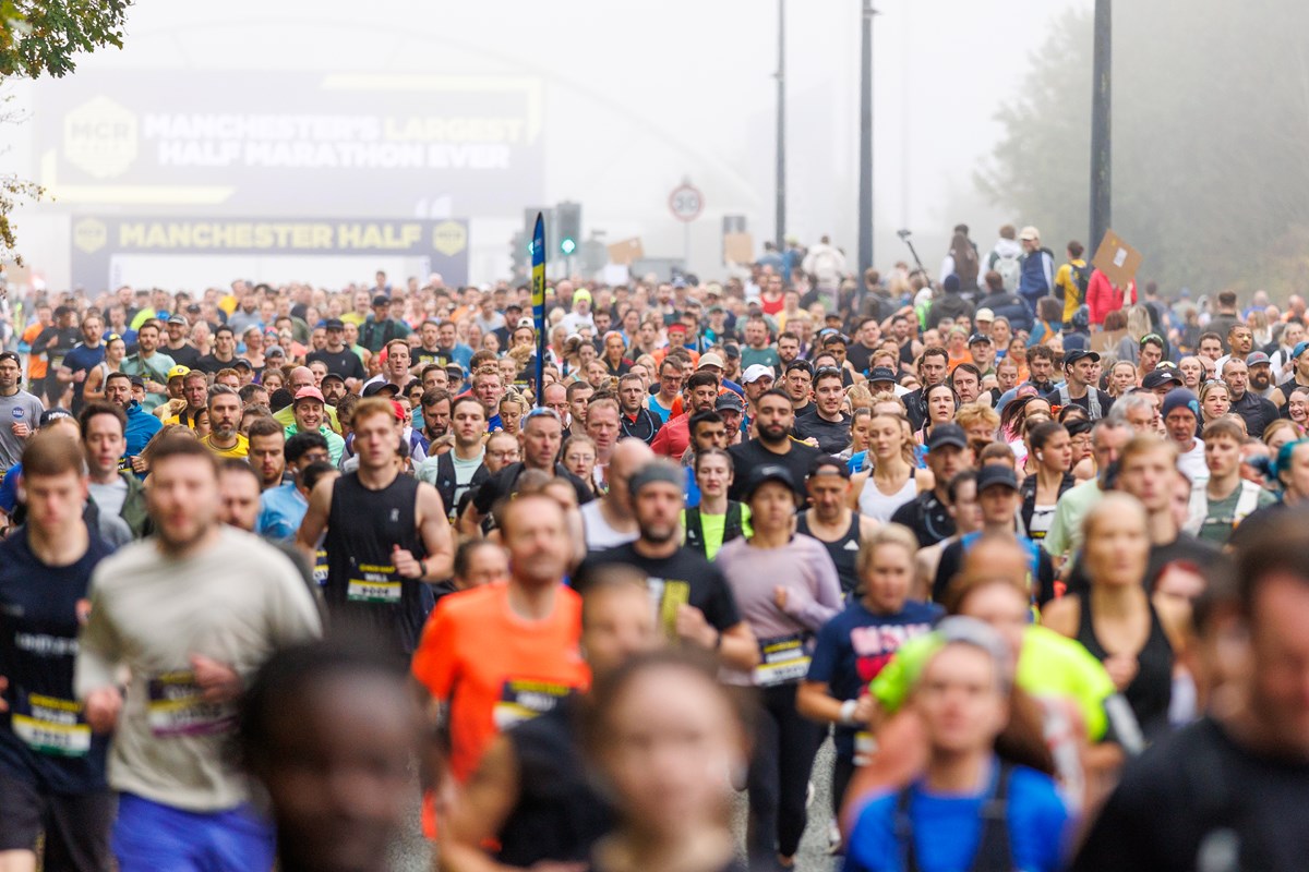 A large crowd of runners setting off from the start line under a banner reading "Manchester Half" and "Manchester's largest half marathon ever".