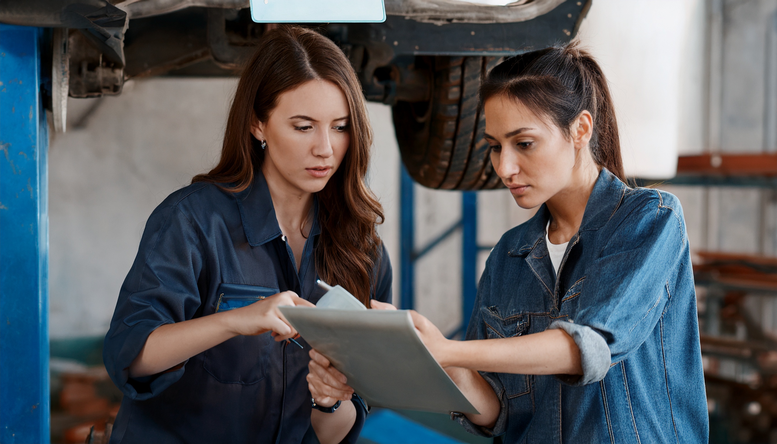 Two female mechanics in workwear look at a clipboard together beneath a car raised on a ramp.