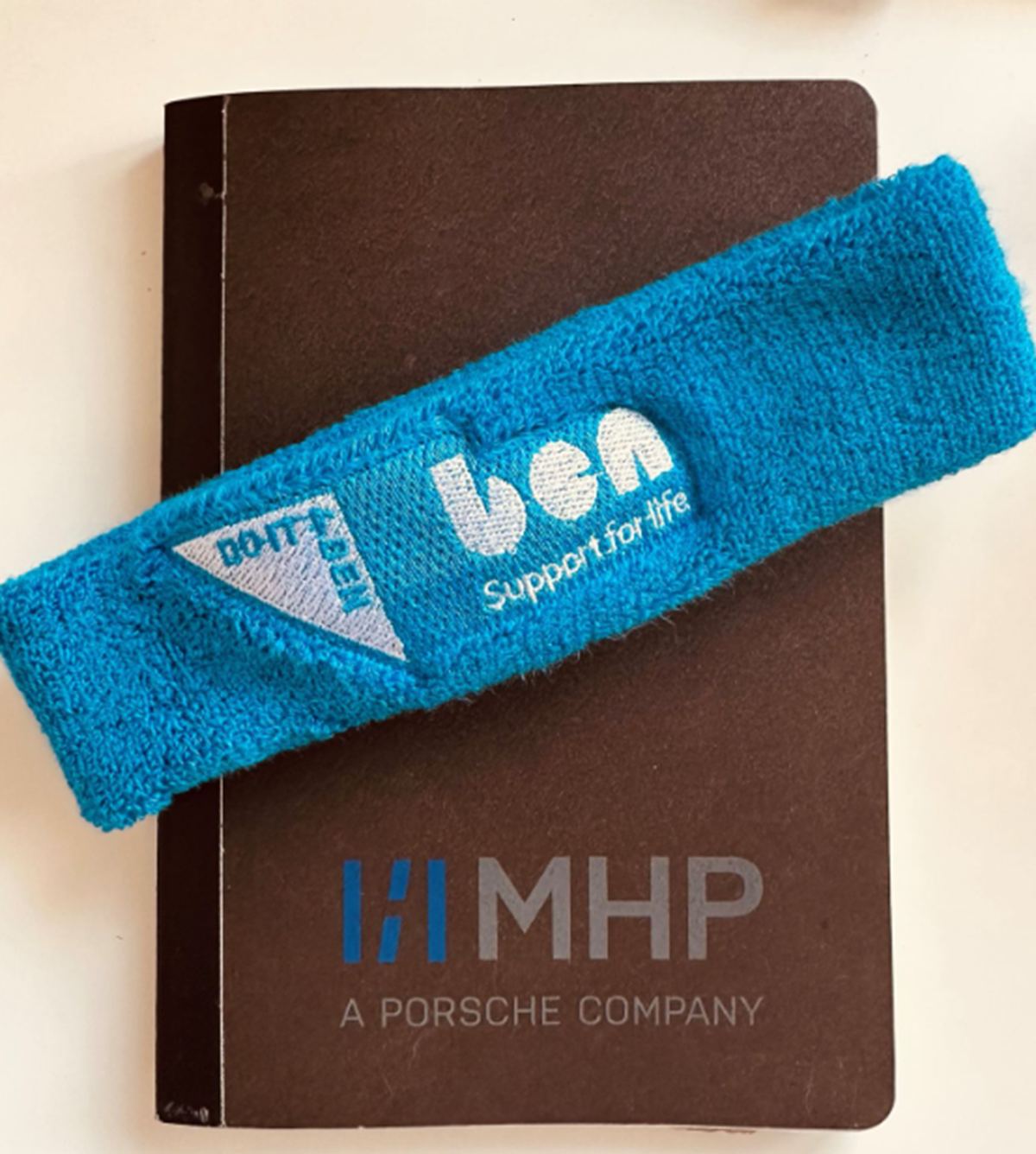 Blue towelling headband embroidered with the Porsche and 'ben - support for life' logos, resting on a brown notebook branded 'MHP, a Porsche company'.