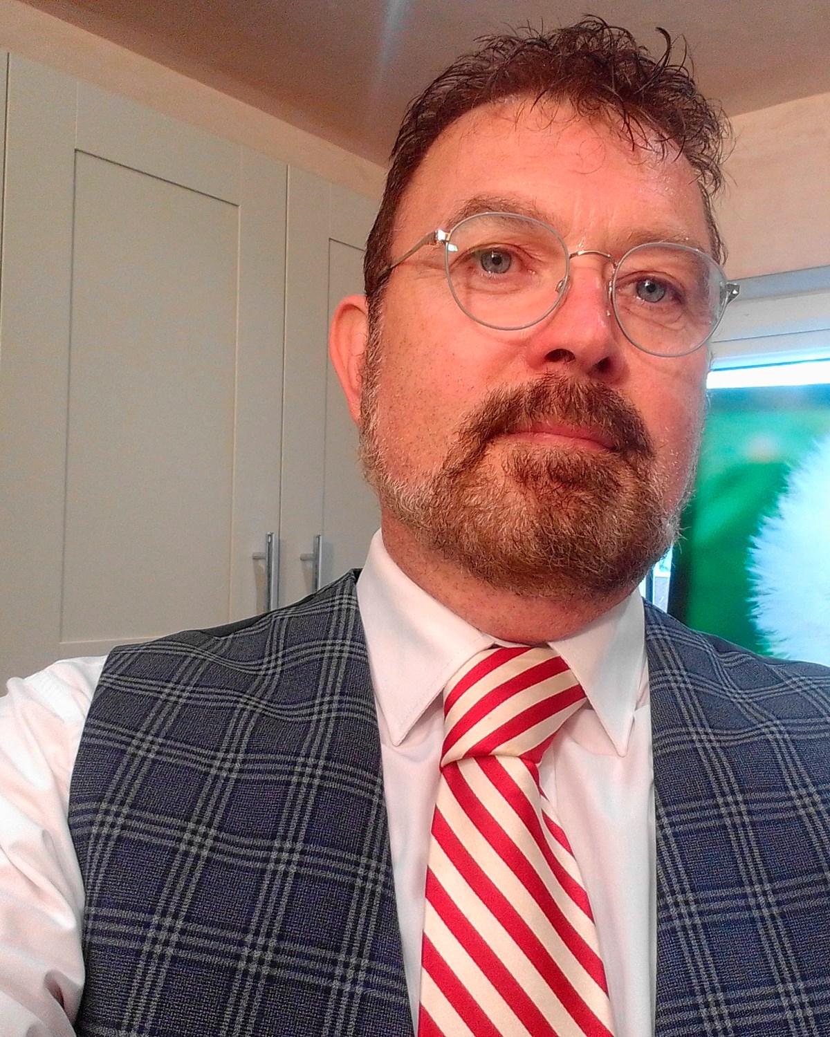 Man with glasses, moustache and beard wearing a checked waistcoat, white shirt and red striped tie.