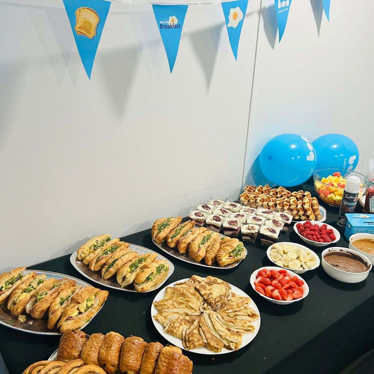 Breakfast buffet of filled croissants, pancakes, waffles, cakes and fruit on a table, under Ben's Big Breakfast bunting and blue Ben balloons.