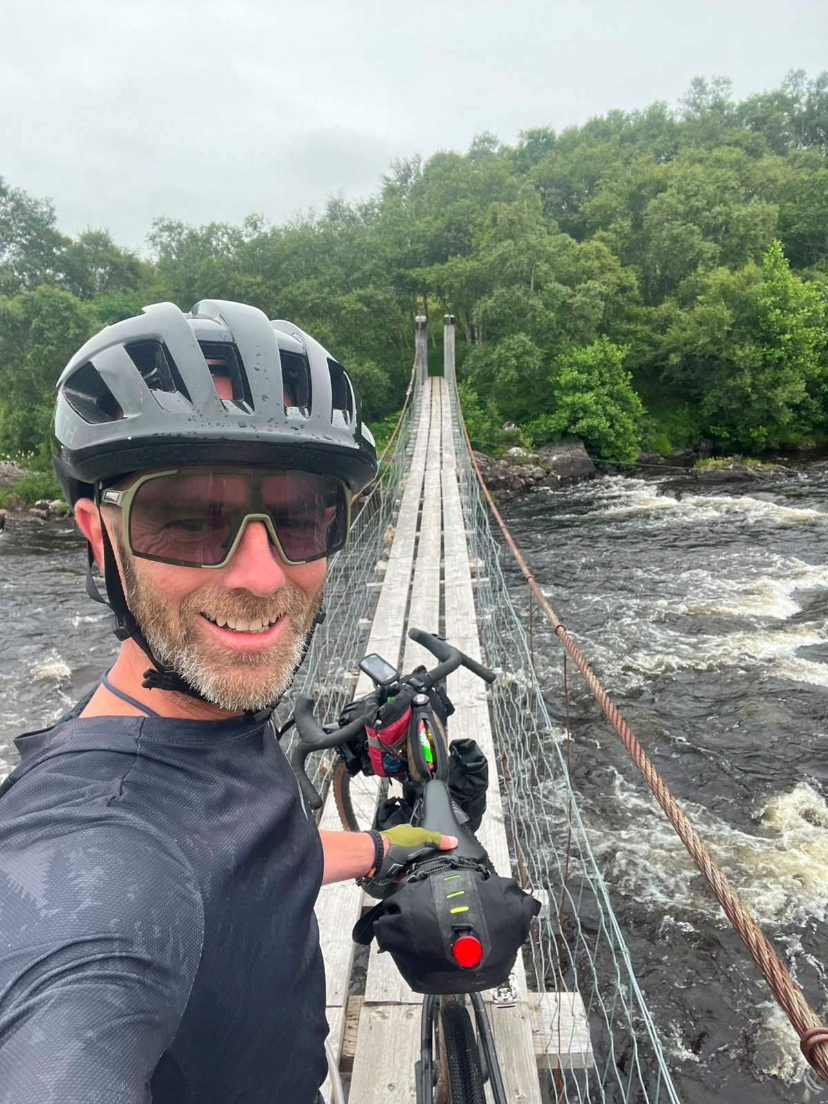 Cyclist in helmet and sunglasses takes a selfie with his loaded bike on a narrow wooden suspension footbridge over a fast-flowing river.