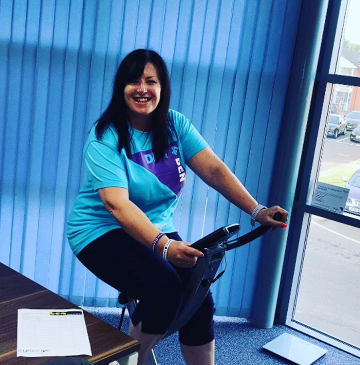 A woman in a Ben T-shirt smiles as she rides an exercise bike indoors beside a window.