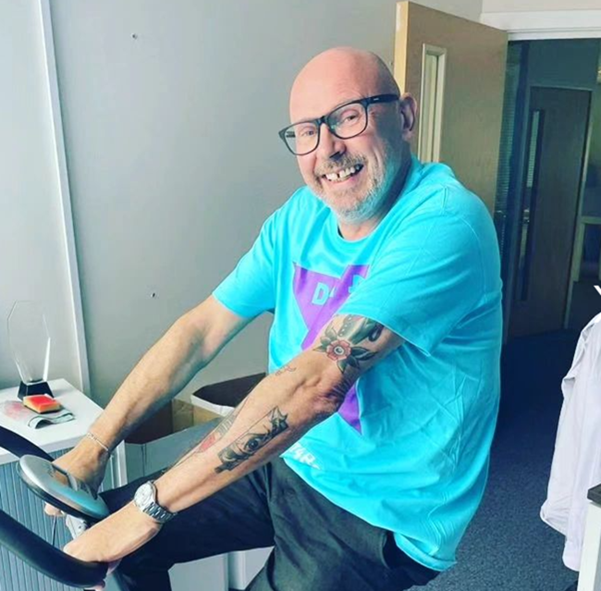 Smiling man in a bright blue T-shirt riding an exercise bike indoors.