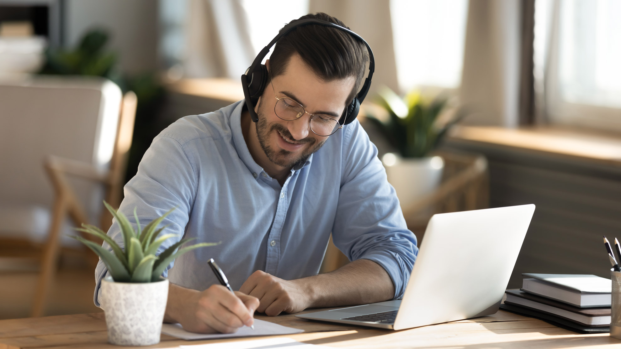 Man wearing a headset smiles as he takes notes at a laptop during an online training session.