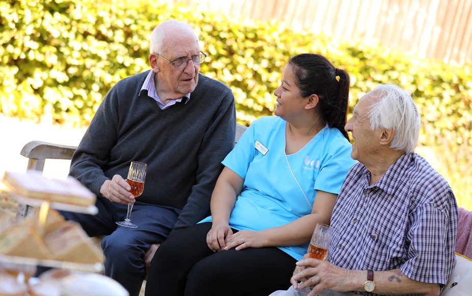Residents are sitting on a bench in the garden, enjoying a glass of wine and chatting with a carer.