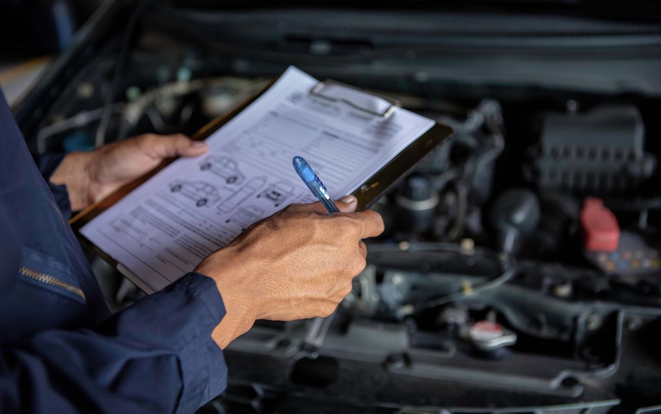 A mechanic servicing a car in a garage - with a clipboard in hand as they check off MOT test.