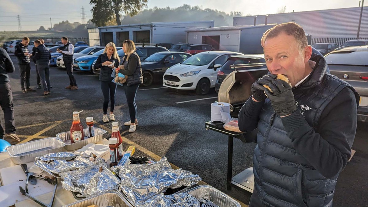 A man eats a bacon roll beside a barbecue at a dealership car park breakfast event, with colleagues chatting nearby.