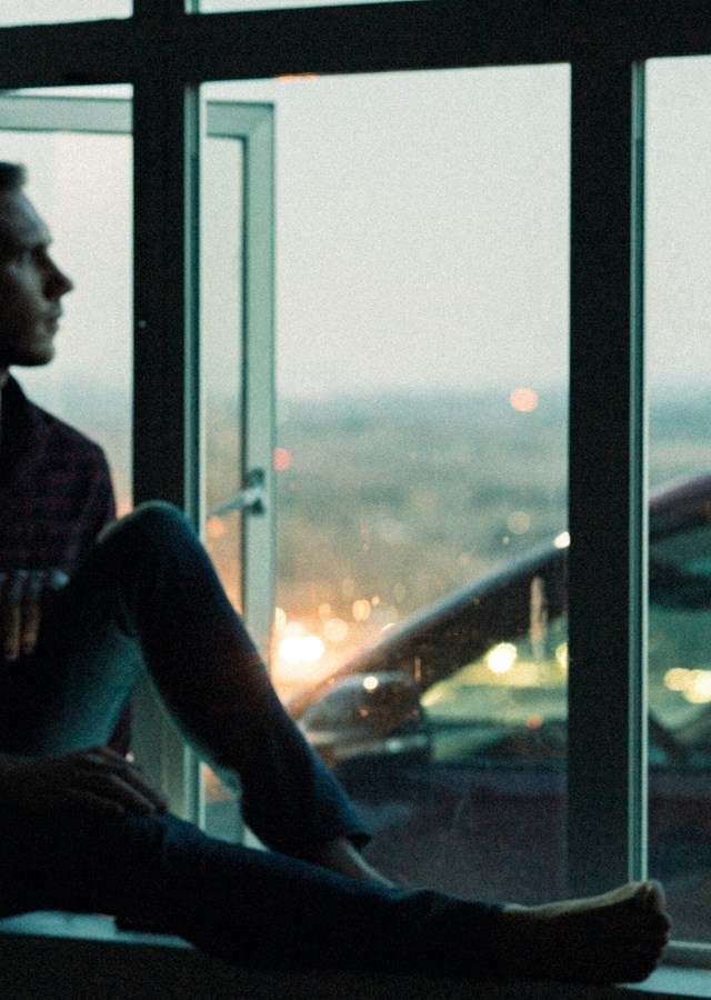 Man sitting on a window sill at dusk staring out of the window