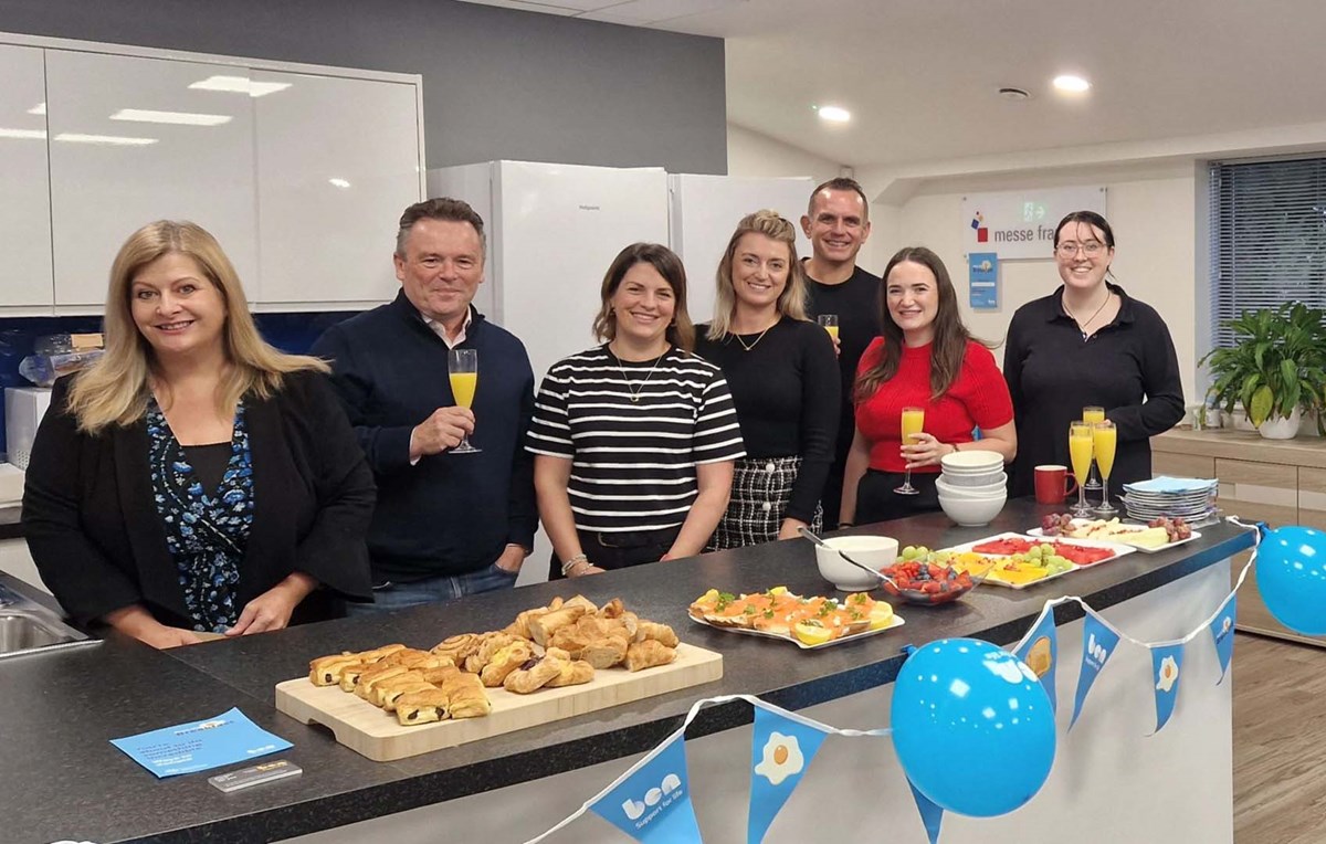 Colleagues gathered around an office kitchen counter with pastries, fruit and orange juice, with blue Ben bunting and balloons, for Ben's Big Breakfast.