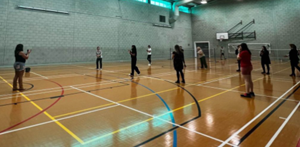 A group of people standing spaced apart in a sports hall, taking part in an indoor exercise session.