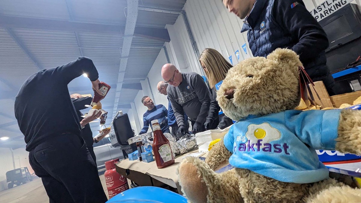 Colleagues serving breakfast rolls and ketchup at a workshop table, with a teddy bear in a 'Big Breakfast' T-shirt in the foreground.