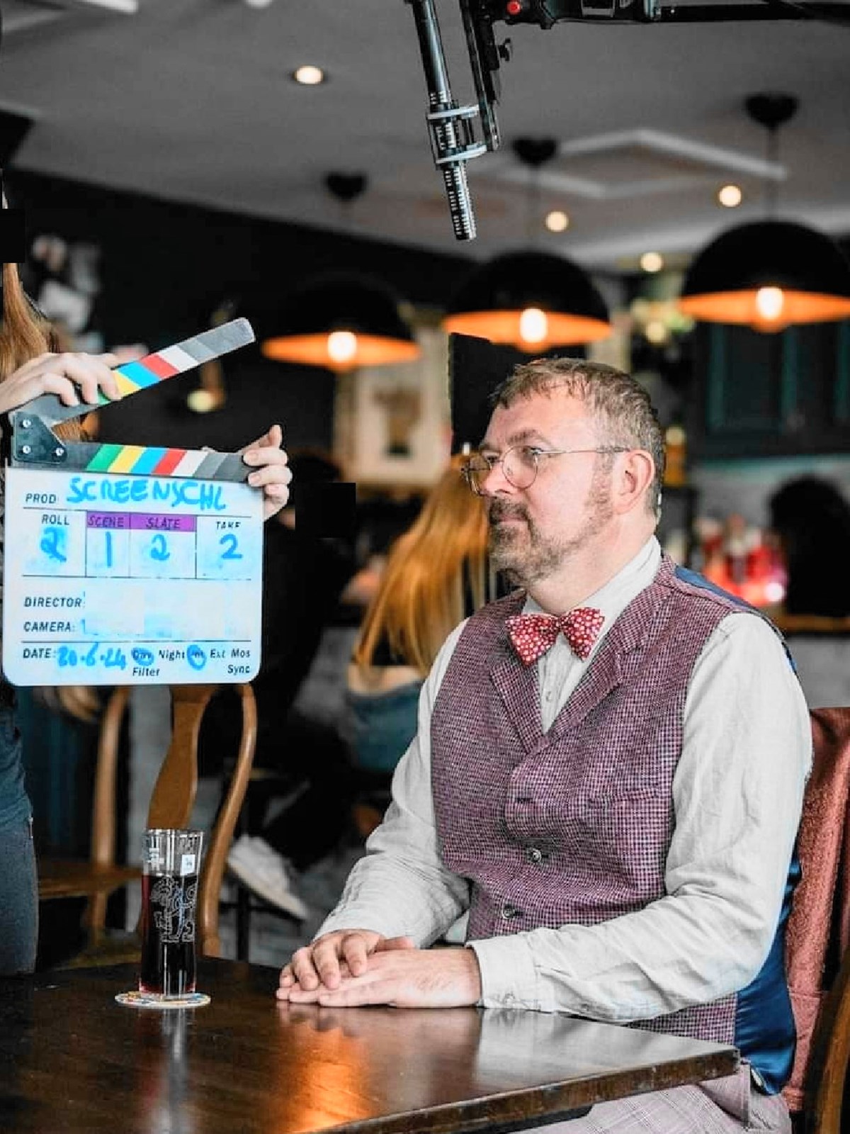 A man in a tweed waistcoat and red bow tie sits at a pub table while a crew member holds a clapperboard beside him during filming.