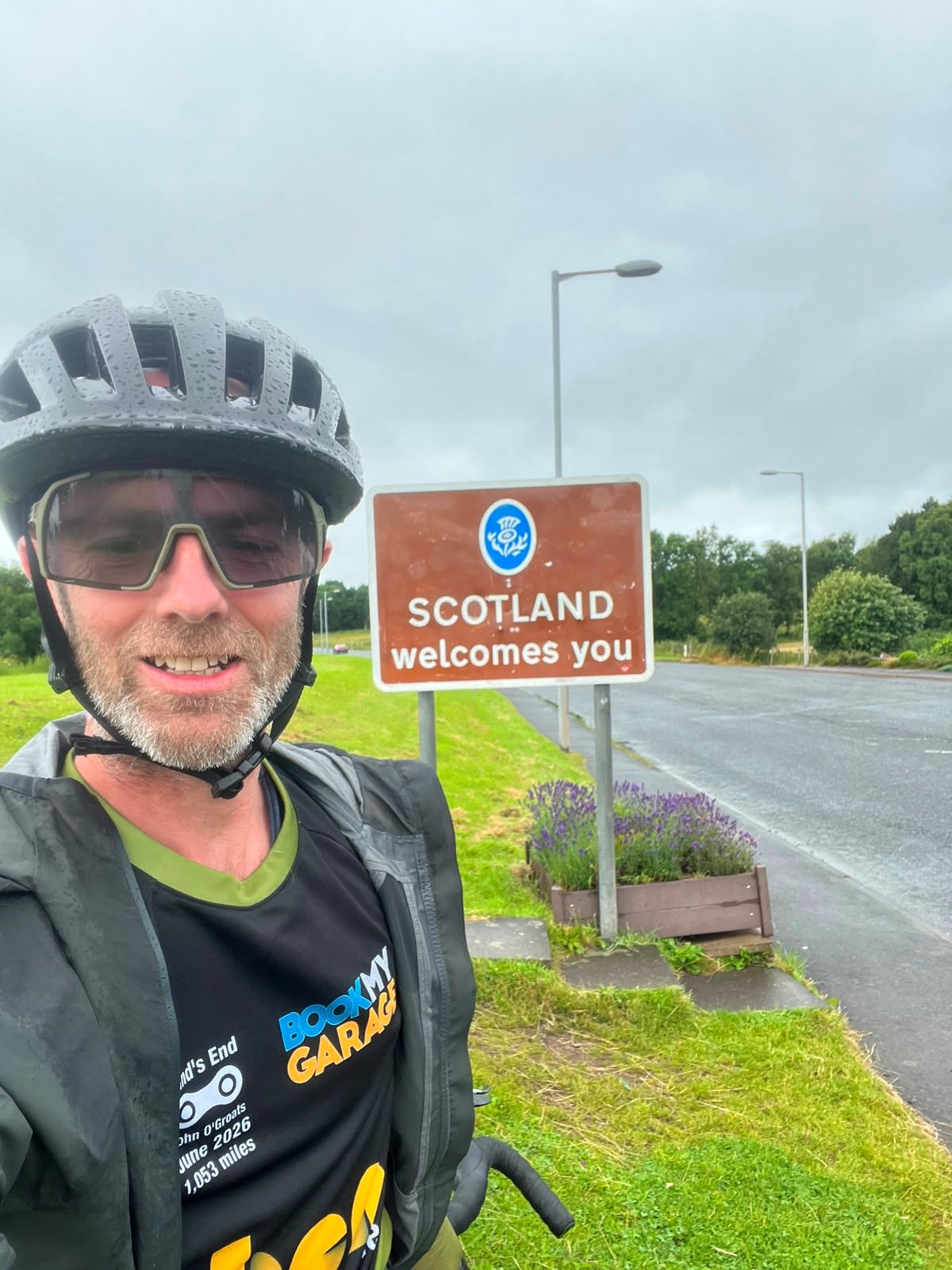 Cyclist in helmet and BookMyGarage jersey takes a selfie beside a roadside sign reading "Scotland welcomes you".