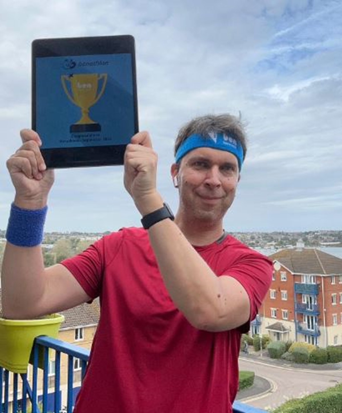 A man in a red t-shirt and blue Benathlon headband holds up a tablet showing a gold trophy certificate on his balcony.