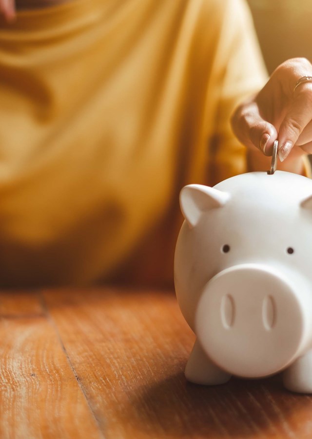 Person saving money and putting coins into a piggy bank with a notebook on the side working out their living costs