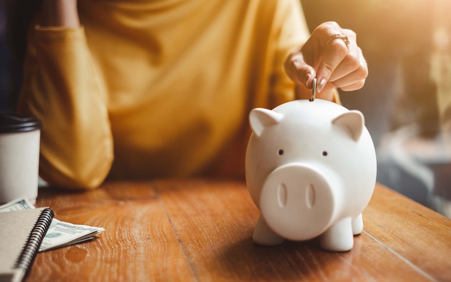 Person saving money and putting coins into a piggy bank with a notebook on the side working out their living costs