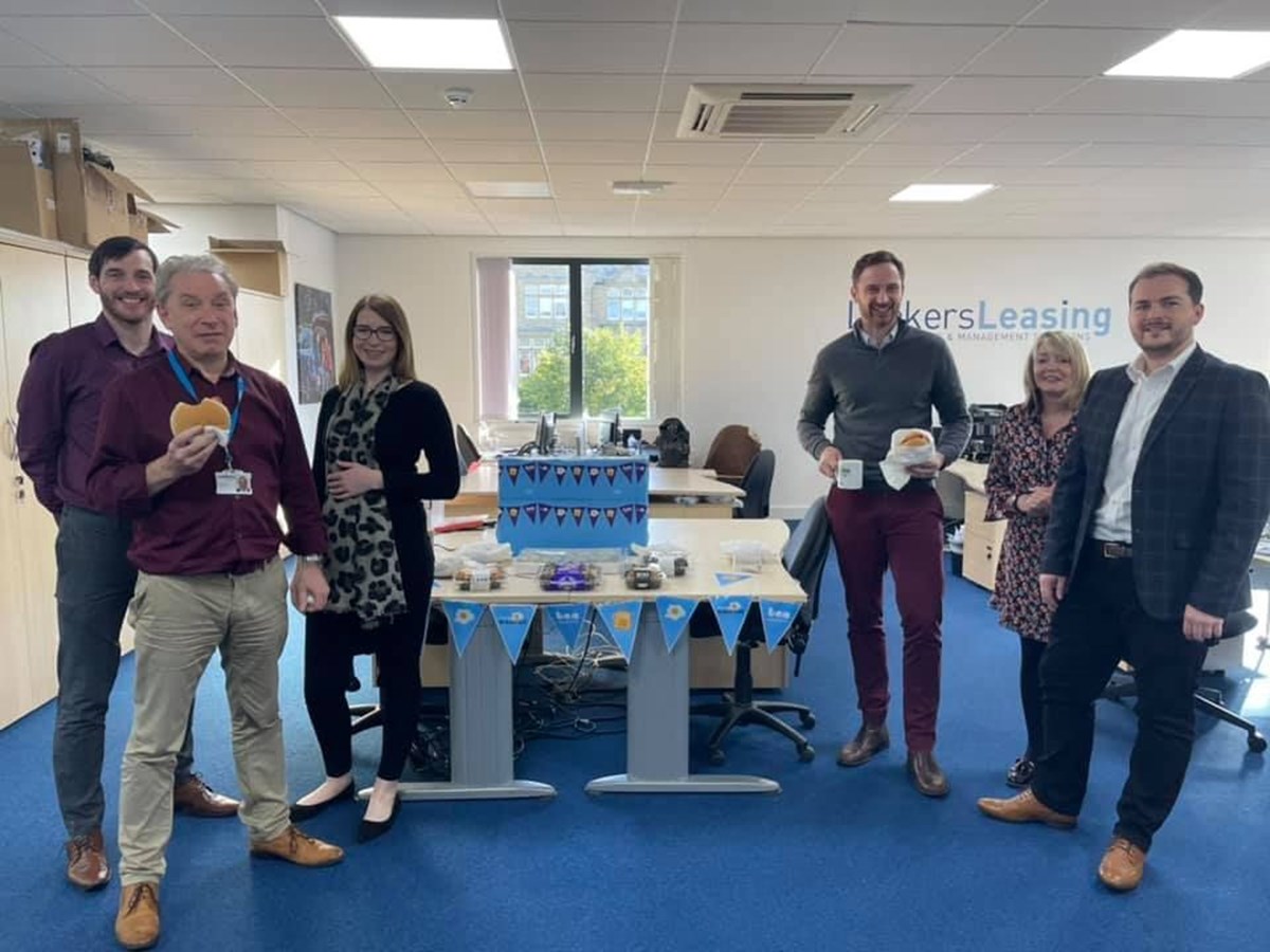 Six office colleagues stand around a desk decorated with blue bunting and cakes, holding breakfast rolls and mugs, in front of a Lookers Leasing wall sign.