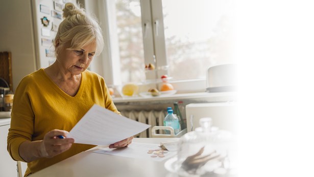 An older woman sits at her kitchen table reading paperwork, with coins beside her.