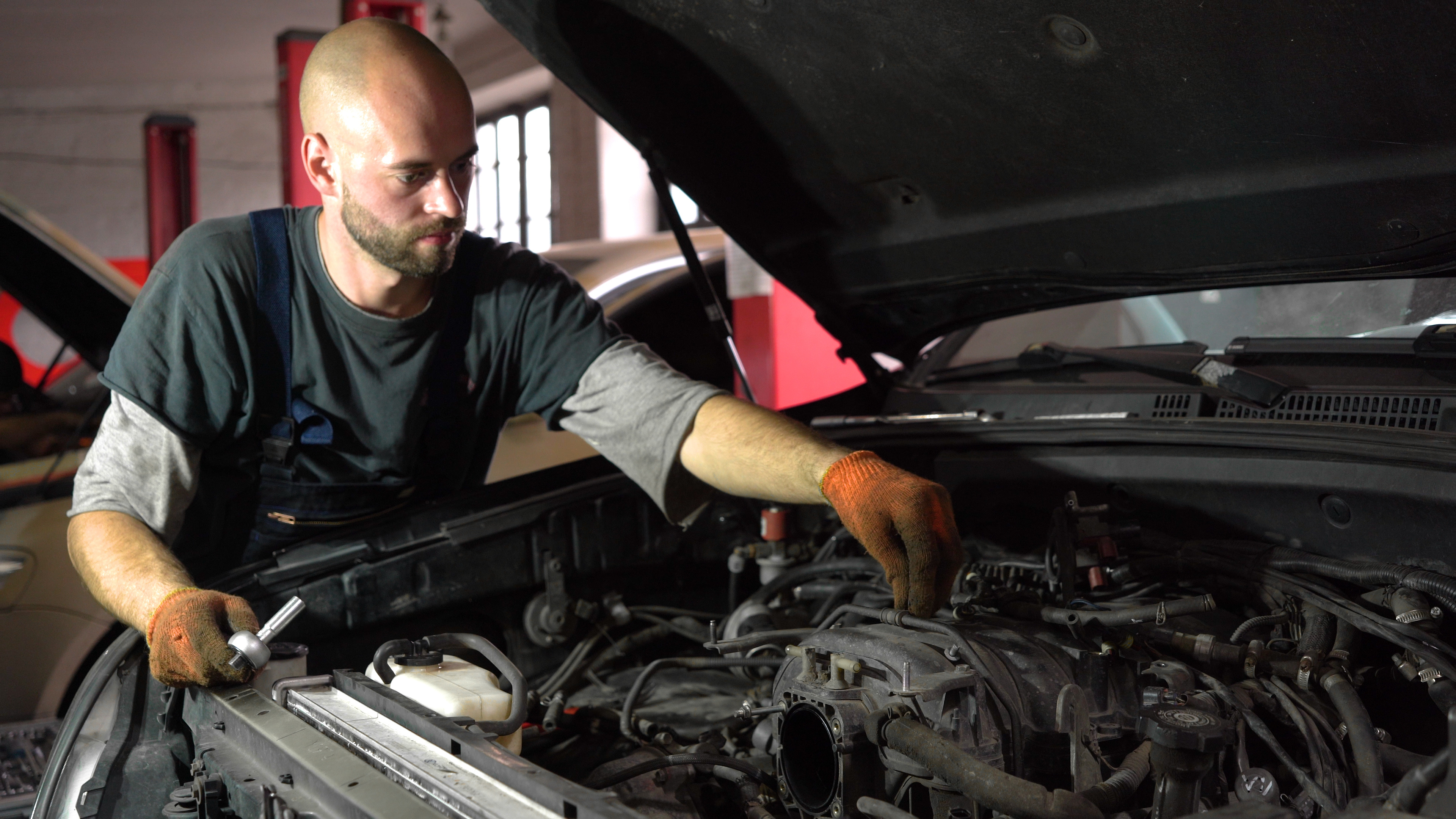 A mechanic in overalls and work gloves leans over the engine bay of a car in a garage.