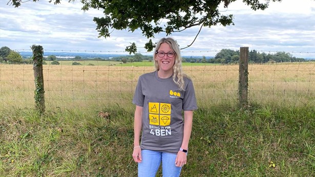 A smiling woman standing in a field beside a wire fence, wearing a grey 'Bring It On 4 Ben' T-shirt.