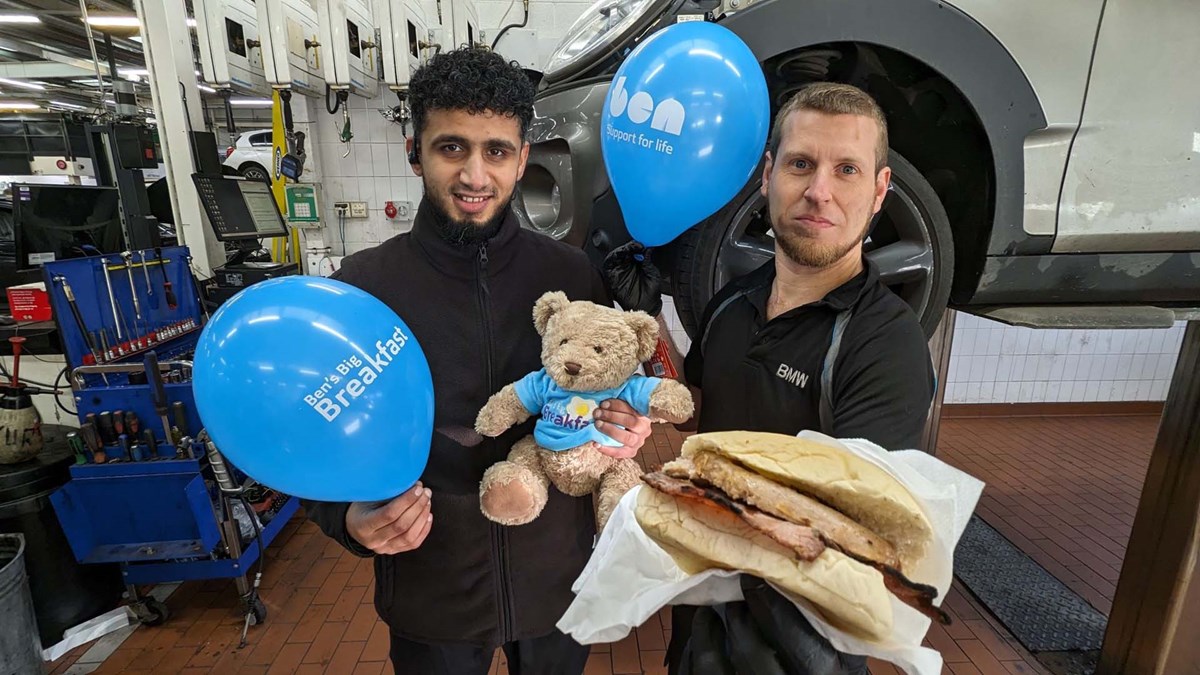 Two workshop colleagues holding blue Ben's Big Breakfast balloons, a Ben teddy bear and a bacon roll in a garage.