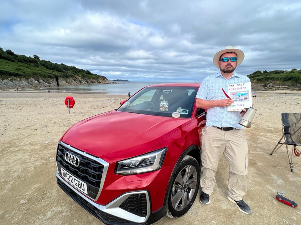 A man in a sun hat and sunglasses stands beside a red Audi on a beach, holding a hand-drawn sign reading "Car Sale Agreement - Sign Here" and a quill pen.