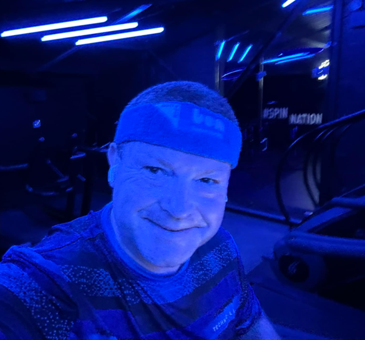 Smiling man wearing a headband takes a selfie in a spin studio lit with blue lights, with '#SPIN NATION' on the wall behind.