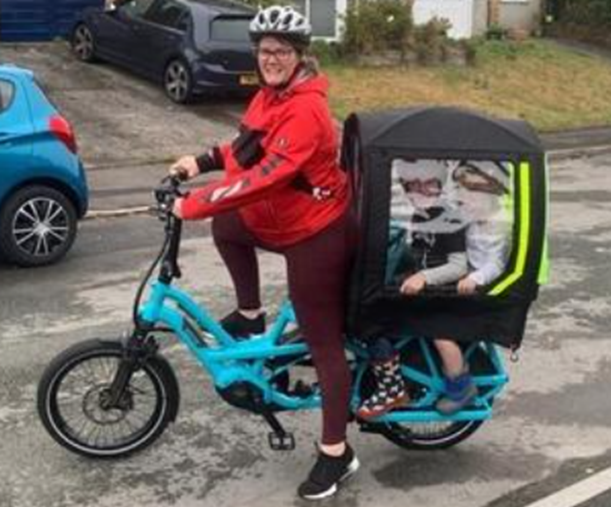 A woman in a red jacket and helmet rides a blue cargo e-bike with two children seated in the covered rear passenger pod.