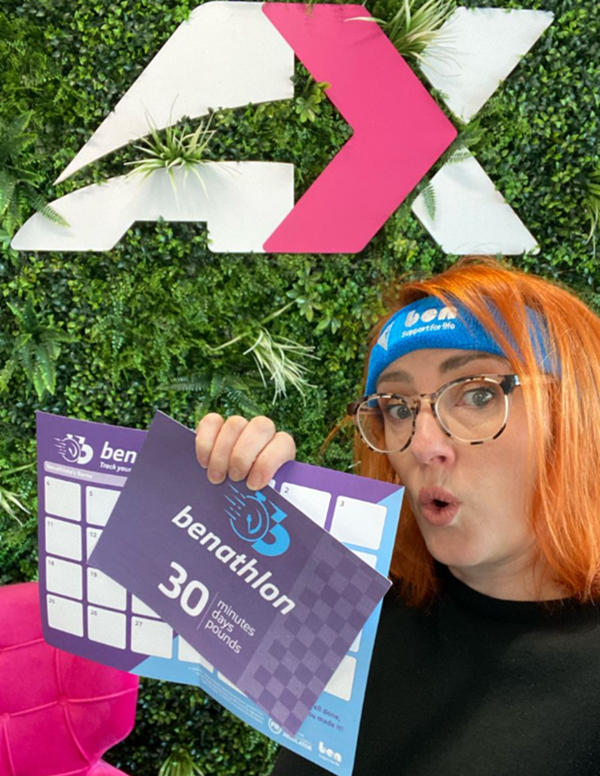 A woman wearing a Ben branded headband holds up Benathlon leaflets and a tracker chart in front of a green foliage wall.