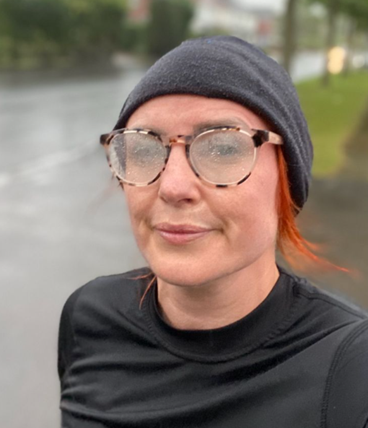A woman in a grey beanie hat, glasses and black running top takes a selfie beside a wet road in the rain.