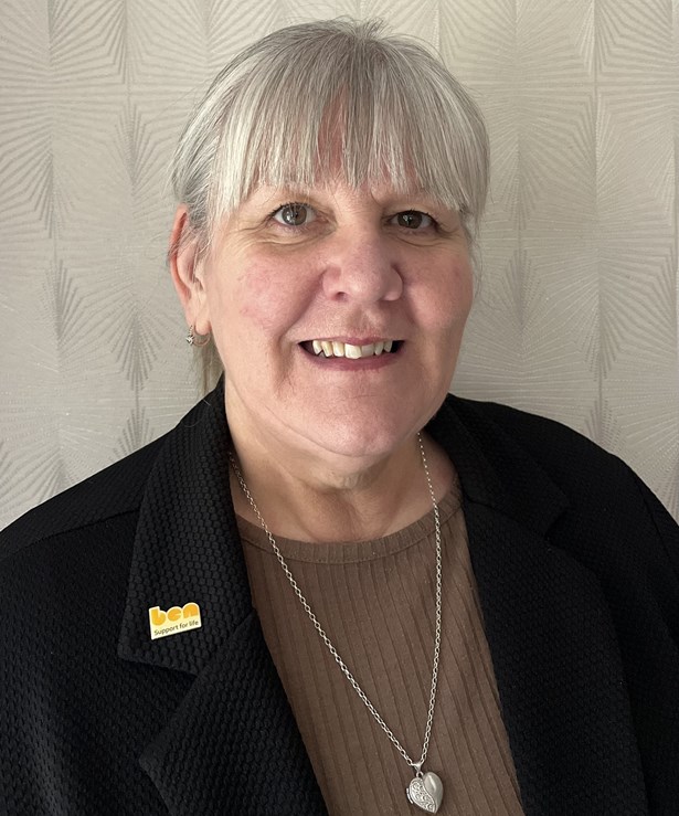 Smiling woman with short grey hair wearing a black jacket with a yellow Ben 'support for life' badge.