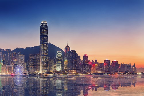 City skyline of illuminated skyscrapers at sunset, reflected in calm harbour water.