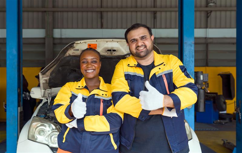 Two mechanics in work uniforms and protective gloves with thumbs up, standing in front of a white car with an open hood.