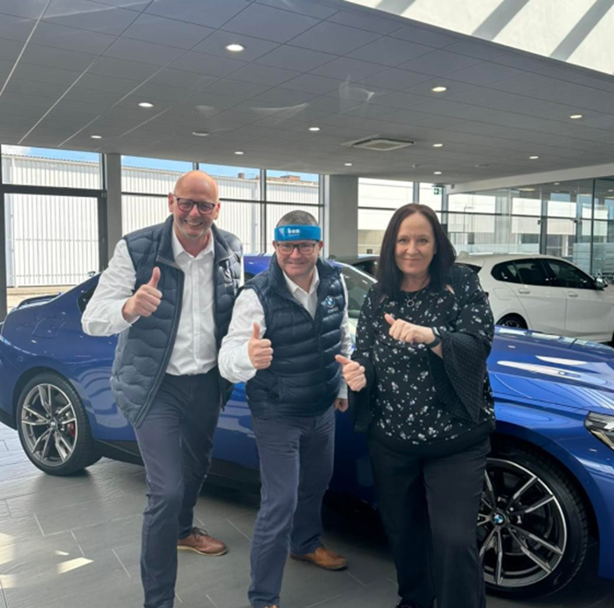 Three car dealership colleagues giving thumbs up in a showroom beside blue and white BMWs, one wearing a Ben headband.