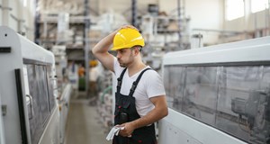 Worker standing in a factory wearing hardhat with one hand on their head while exhaling deeply