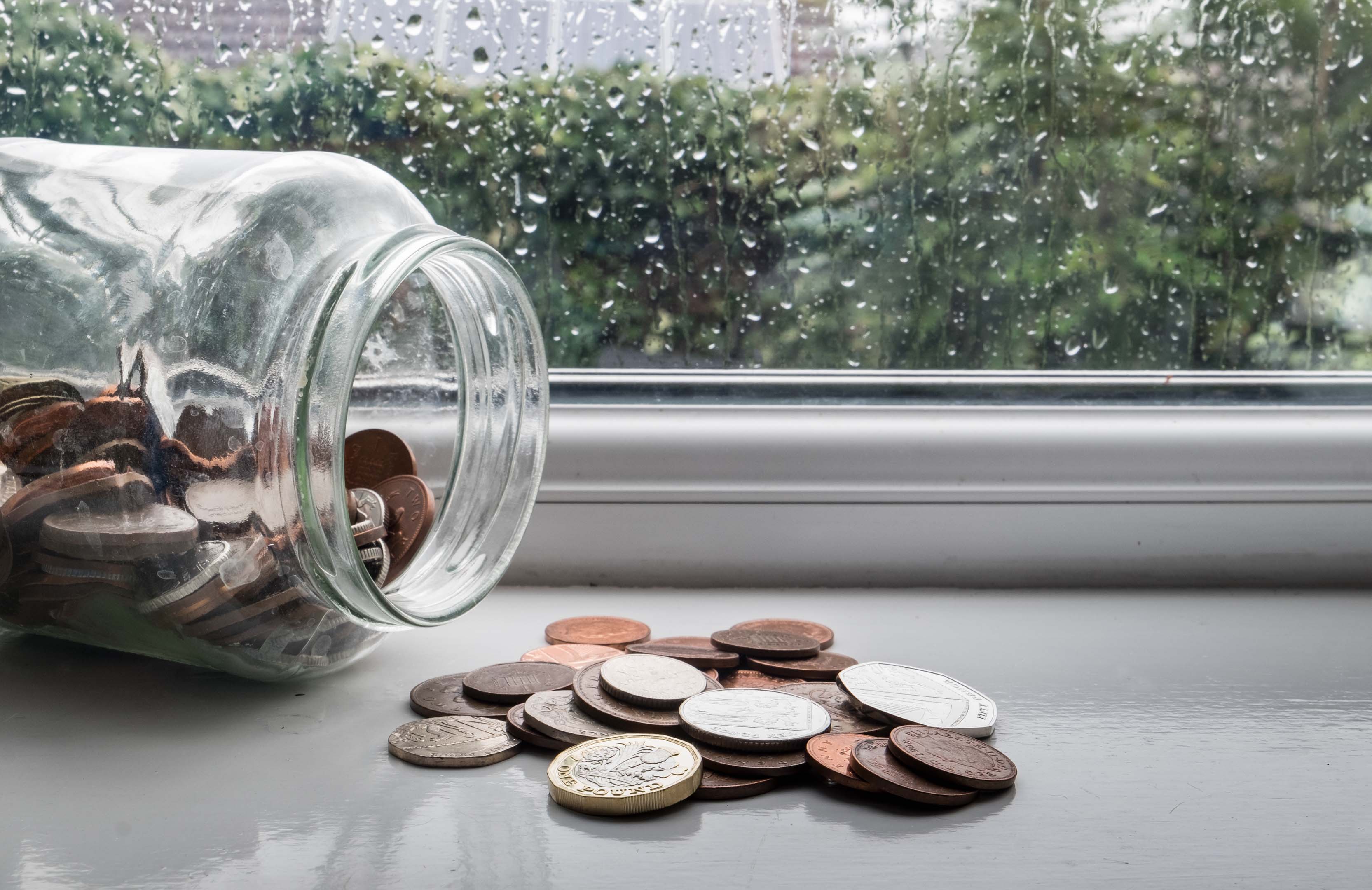 A jar full of money on its side with some of the coins spilled out on a windowsill on a rainy day