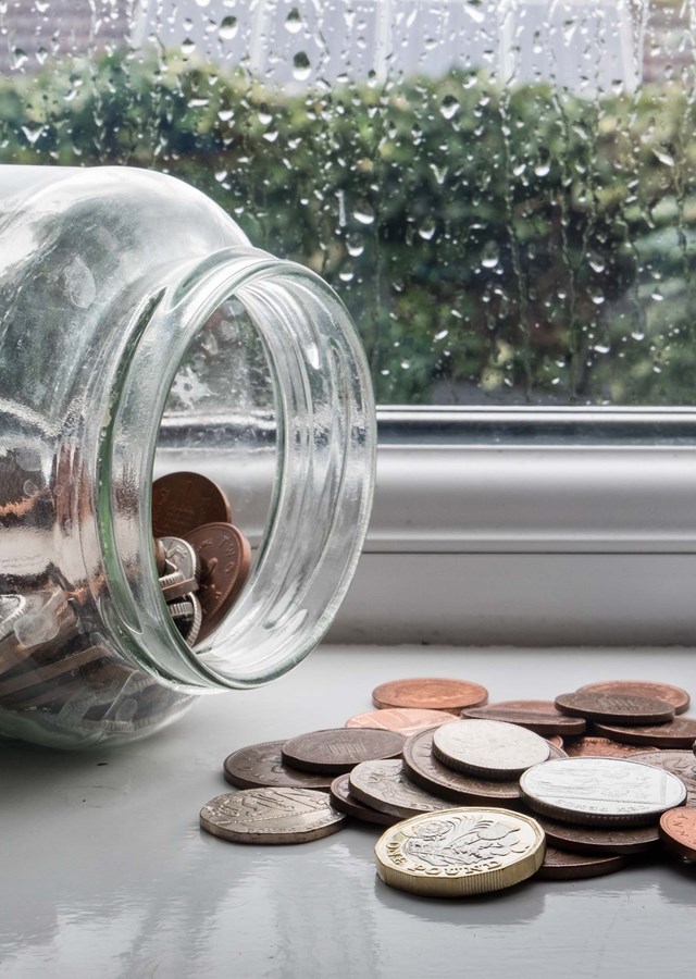 A jar full of money on its side with some of the coins spilled out on a windowsill on a rainy day