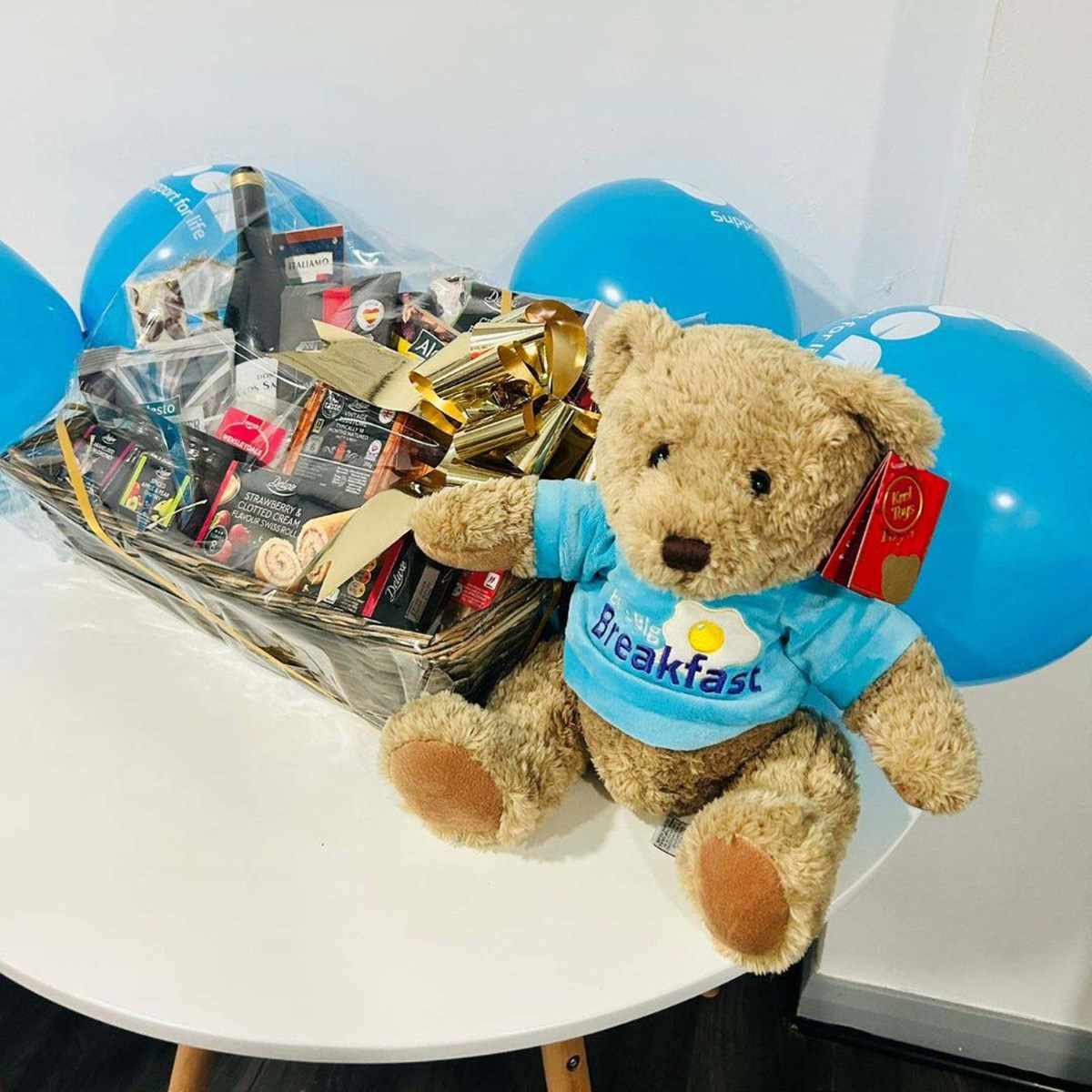 Teddy bear in a 'Ben's Big Breakfast' T-shirt beside a raffle hamper of food and wine, with blue Ben balloons behind.