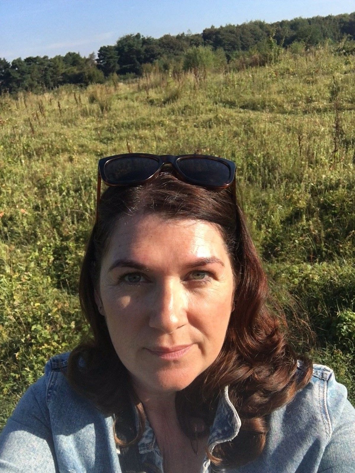 Selfie of a woman with sunglasses on her head, standing in a grassy field with trees behind her